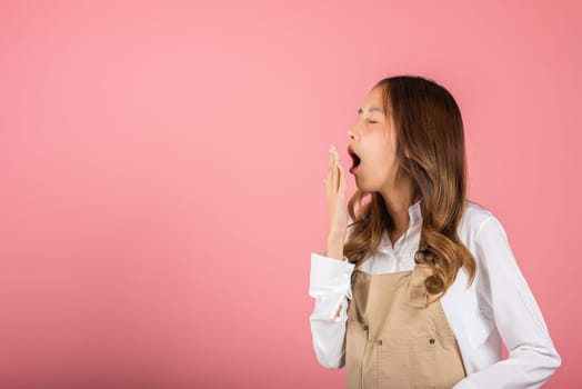 Portrait Asian beautiful young woman wearing overalls emotions tired and sleepy her yawning covering mouth open by hand, studio shot isolated on pink background, Female attractive insomnia boring