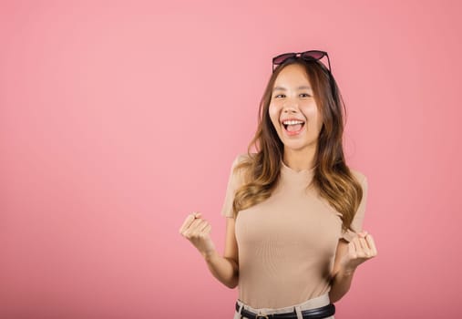 Happy Asian portrait beautiful cute young woman wear glasses makes raised hand up celebrating her winning success gesture, studio shot isolated pink background, Female excited say yes with copy space