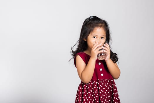 Little cute kid girl 3-4 years old smile drinking fresh water from glass in studio shot isolated on white background, Asian children preschool, Daily life health