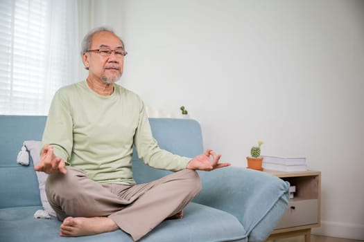 Lifestyle calm senior man lotus pose doing yoga for mental balance breathing air relaxing on sofa at home, Asian elderly old man sitting practise yoga and meditation in lotus position and closed eyes