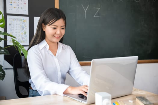 Back to school. Smiling female using computer sitting at school table, Portrait of young woman teacher with laptop at desk in classroom, Online education and learning concept