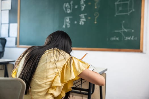 Education. Back view of school girl on lesson in classroom write hardworking on blackboard, primary child is sitting lessons at table in school writing or drawing in notebook, Back to school concept