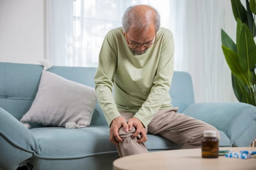 Old man suffering from knee joint pain on sofa living room, bone pain in elderly at home, senior man knee problem painful, unhappy old age hand holding on knee pain after tendon surgery