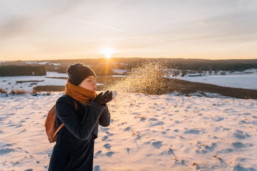 A woman in a coat wrapped in a scarf blows snow off her hands.