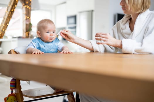 Mother wearing cosy bathrope spoon feeding her baby boy child in baby high chair with fruit puree at dinning table at home. Baby solid food introduction concept.