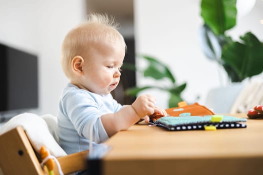 Happy infant sitting at dining table and playing with his toy in traditional scandinavian designer wooden high chair in modern bright atic home. Cute baby playing with toys.