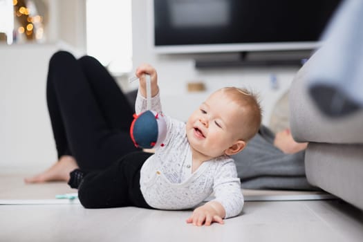 Happy family moments. Mother lying comfortably on children's mat watching and suppervising her baby boy playinghis in living room. Positive human emotions, feelings, joy.