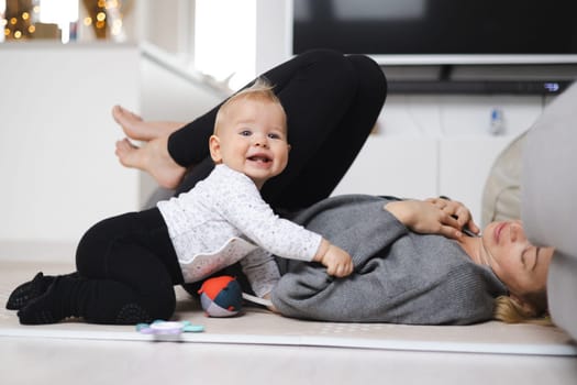 Happy family moments. Mother lying comfortably on children's mat playing with her baby boy watching and suppervising his first steps. Positive human emotions, feelings, joy