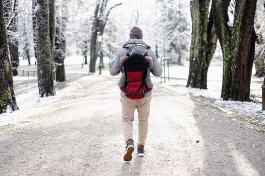 Rear view of sporty father carrying his infant son wearing winter jumpsuit and cap in backpack carrier walking in city park in winter.