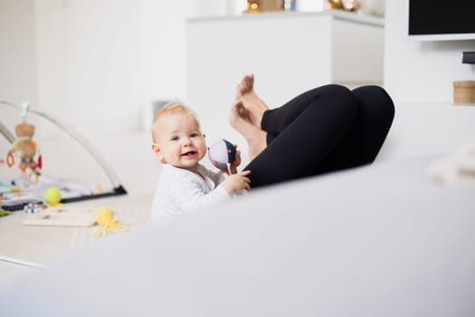 Happy family moments. Mother lying comfortably on children's mat playing with her baby boy watching and suppervising his first steps. Positive human emotions, feelings, joy