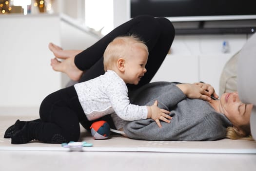 Happy family moments. Mother lying comfortably on children's mat playing with her baby boy watching and suppervising his first steps. Positive human emotions, feelings, joy