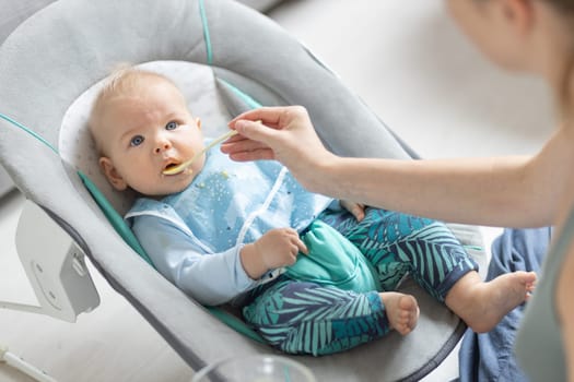Mother spoon feeding her baby boy child in baby chair with fruit puree. Baby solid food introduction concept