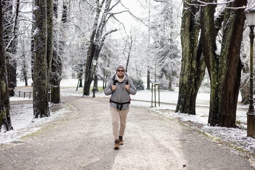 Sporty father carrying his infant son wearing winter jumpsuit and cap in backpack carrier walking in city park in winter.