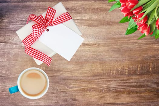 Coffee mug, present box with blank paper tag and tulips on rustic wooden table. Top view. Breakfast on Valentine, Mother or Womens Day. Copy space. Card concept.