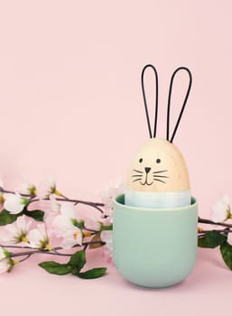Cute easter bunny or rabbit with a smiling egg face in a cup, spring holiday greeting card with cherry blossoms