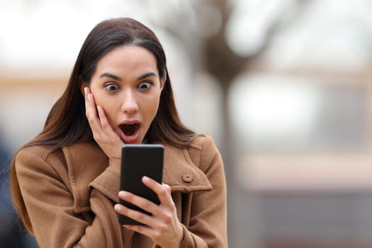 Amazed woman in winter checking cell phone in the street