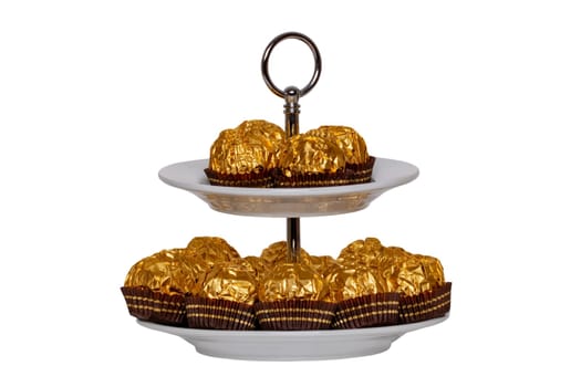 Pastries, desserts and sweets. Chocolate candies wrapped in golden foil on a two-storey etagere isolated on a white background. Clipping path. Decoration element for greeting card, poster or invitation. Macro.