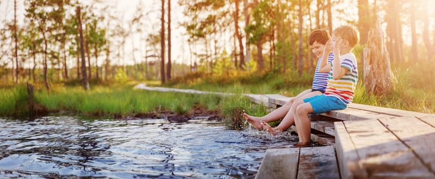 Two boys sitting on the pier of the swamp lake and making splashes by legs. Concept of the family vacation and friendship.