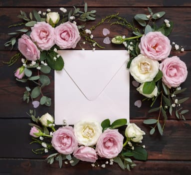 Square Envelope between pink and cream roses on brown wood top view, wedding mockup. Romantic scene with envelope and pastel flowers flat lay. Valentines, Spring or Mothers day concept