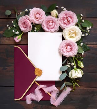 Card and red envelope between pink and cream roses on brown wood top view, wedding mockup. Romantic scene with cards, envelope and pastel flowers flat lay. Valentines, Spring or Mothers day concept