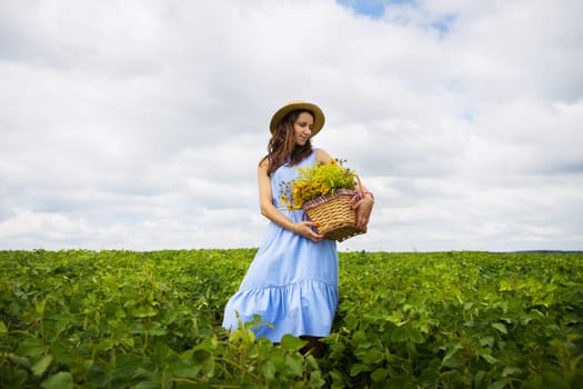 girl in a hat stands on a green field with a basket of flowers.