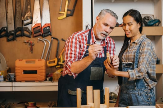 Craftsmen senior man teaching woman apprentice at woodshop, Carpenter training wood for female standing at wood table in workshop, National Carpenters Day
