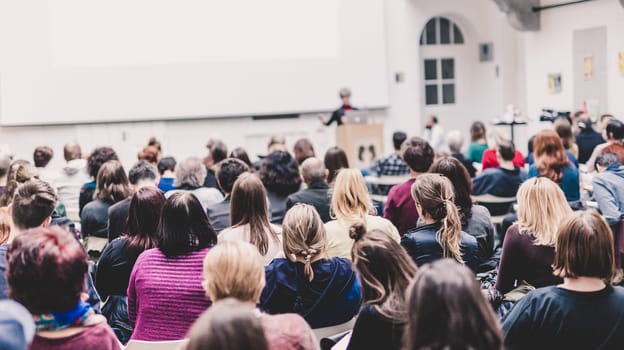 Business and entrepreneurship symposium. Female speaker giving a talk at business meeting. Audience in conference hall. Rear view of unrecognized participant in audience. Copy space on whitescreen.