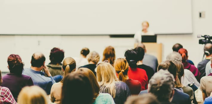 Business and entrepreneurship symposium. Female speaker giving a talk at business meeting. Audience in conference hall. Rear view of unrecognized participant in audience. Copy space on whitescreen.