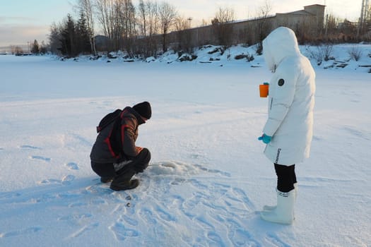 Logmozero, Karelia, Russia, January 08, 2022. Fisherman and fisherwoman on winter fishing. A man sits near a hole in a frozen lake and waits for a bite. Snow on ice. Bottom fishing for perch.