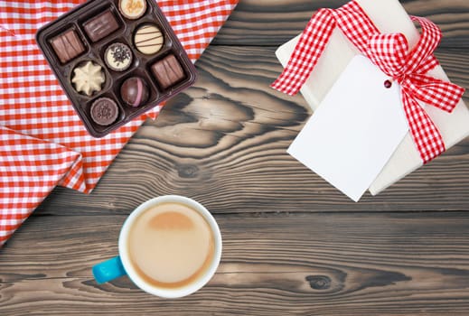 Coffee mug, present box with blank paper tag and box of fine chocolate candies on rustic wooden table. Top view. Breakfast on Valentine, Mother or Womens Day. Copy space. Card concept.