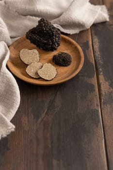 Whole and sliced black truffles mushroom on wooden plate on dark brown table near grey napkin, close up, copy space. Culinary delight ingredient.