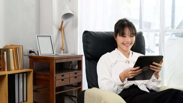 Beautiful asian woman reading or watching content on digital tablet in a bright living room.