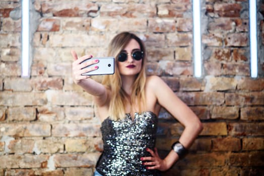 Young sexy woman posing in nightclub and taking selfie with her smartphone. Glamour stylish beautiful girl having fun in front of disco brick wall