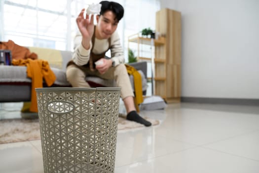 Young man throwing a crumpled paper into a trash bin while tidying up his living room.