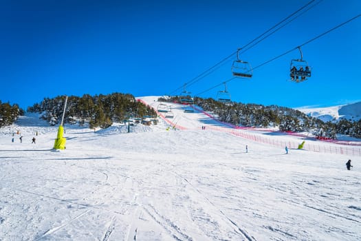 People riding up on a ski chair lift. People skiing and snowboarding down on the ski slope. Winter holidays in Andorra, El Tarter, Pyrenees Mountains