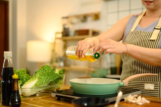 Middle aged woman in apron preparing delicious home cooked meal, adding oil to a frying pan.