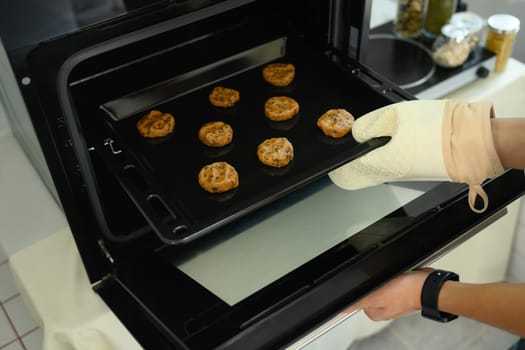 Baker wearing an oven mitt placing a tray of chocolate chip cookie dough into the oven.
