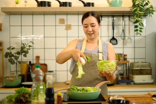 Smiling housewife preparing healthy meal in a modern kitchen adding fresh green vegetables to a pan.