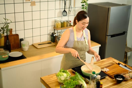 Smiling middle aged woman stirring ingredients in frying pan while preparing meal in cozy kitchen.