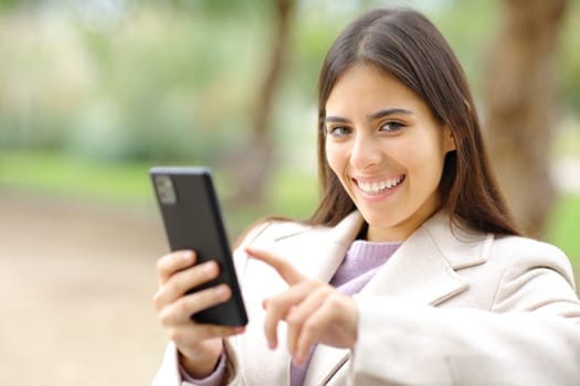 Woman in winter looks at you holding phone in a park
