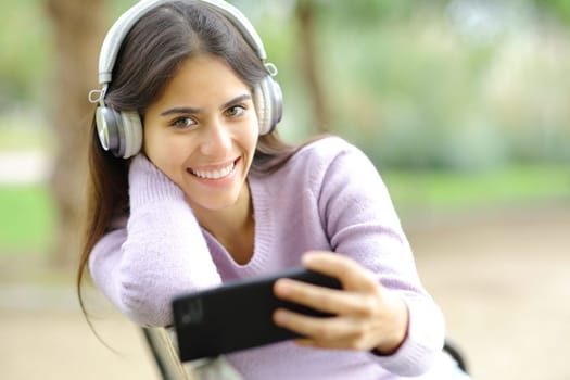 Happy woman listening to music looking at you in a park