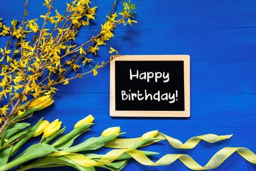 Blackboard With English Text Happy Birthday. Yellow Spring Flowers Like Tulip And Branches. Festive Decoration With Ribbon. Blue Wooden Background