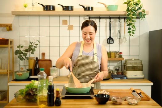 Smiling middle aged woman stirring ingredients in frying pan while preparing meal in cozy kitchen.