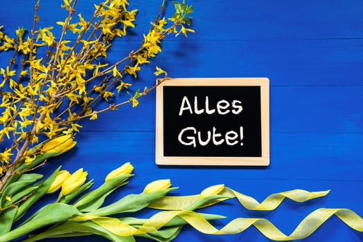 Blackboard With German Text Alles Gute Means Best Wishes. Yellow Spring Flowers Like Tulip And Branches. Festive Decoration With Ribbon. Blue Wooden Background