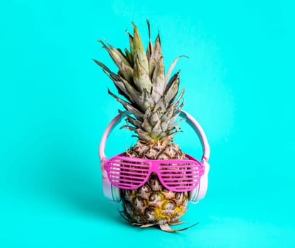 Fashionable  trendy pineapple fruit with headphones and sun glasses listen to the music over bright pastel cyan background. Cool hipster ananas with funny face on blue. Summer holiday vacation and tropical beach party concept.