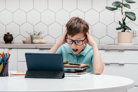 Portrait of young preteen surprised boy wearing glasses, sitting at desk, looking with open mouth at notebook near tablet, touching head, having online lesson. Technology, e-learning, overemotive.