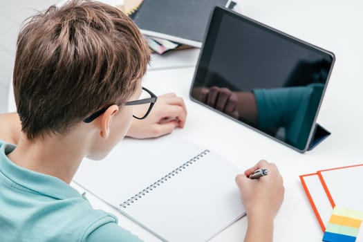 Top view of young preteen boy wearing blue T-shirt, glasses, sitting at white desk near notepads in front of tablet, having online lesson, writing on notebook with left hand. E-learning, technology.
