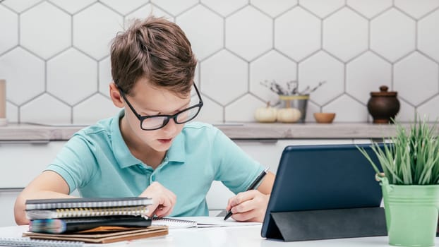Portrait of preteen concentrated boy pupil wearing blue T-shirt, glasses, sitting at desk near notebooks, tablet, doing homework with left hand, writing on notebook. Distant learning, education.