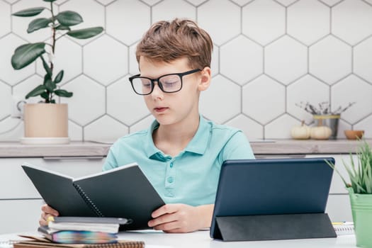 Portrait of young preteen serious boy wearing blue T-shirt, glasses, sitting at desk near notepads, looking at book near tablet, houseplant, having online lesson, reading. E-learning, technology.
