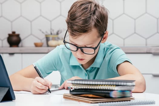 Portrait of preteen serious boy wearing blue T-shirt, sitting at desk near notebooks, tablet, having online lesson, making notes, writing on notebook, holding pen with left hand. Distant learning.